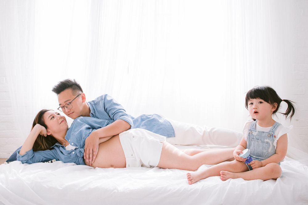 Family in casual attire relaxes together on white bedding, with small child sitting at their feet.