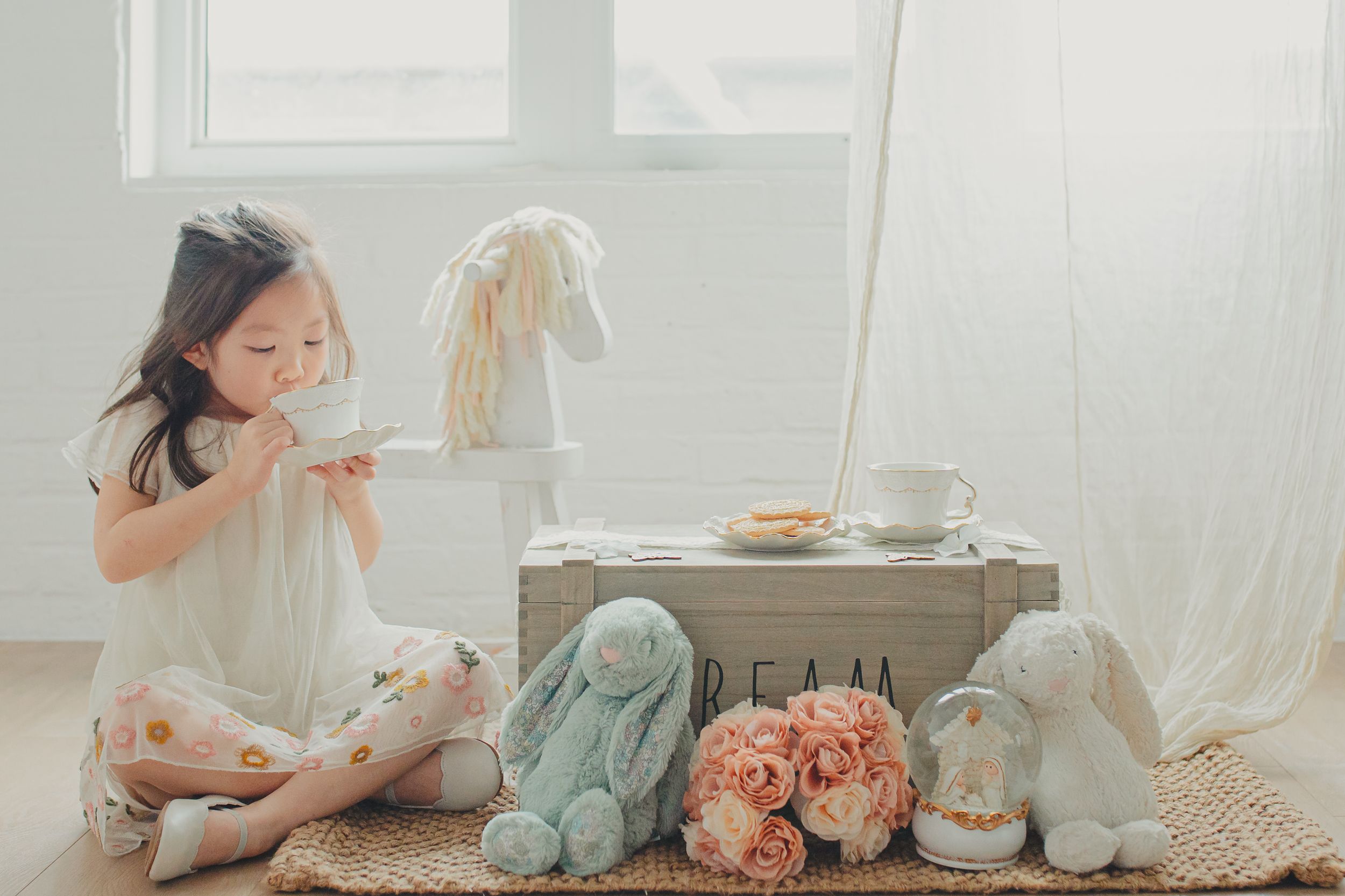 Soft pastel scene with a child playing with dolls and stuffed toys surrounded by pink roses and vintage decor.