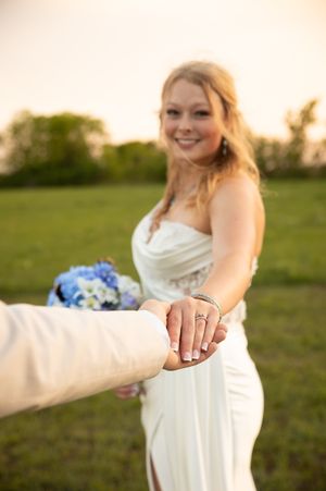 A young person in a white wedding dress holds hands with someone in a field at sunset during a golden hour photoshoot.