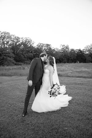A romantic black and white wedding portrait showing a couple sharing an intimate moment in a meadow with trees in background.