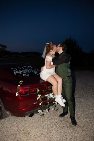 A couple shares a romantic moment on a decorated red car during a nighttime wedding celebration.