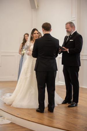 Wedding ceremony in progress inside an elegant white-walled room with hardwood floors.