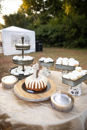 A rustic dessert table with a bundt cake, tiered serving stands, and white pastries arranged on a beige tablecloth outdoors.