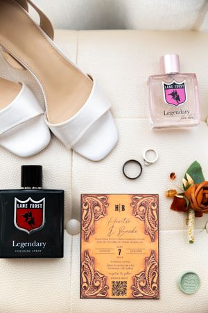 Flat lay of white bridal shoes, wedding rings, perfume bottles, and vintage-style invitation cards on a light background.