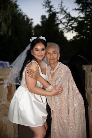 A bride in a white dress and grandmother in a sparkly champagne dress share tender moments at an outdoor evening wedding.