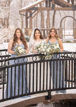 Three women in formal attire stand on a snowy bridge holding winter wedding bouquets.