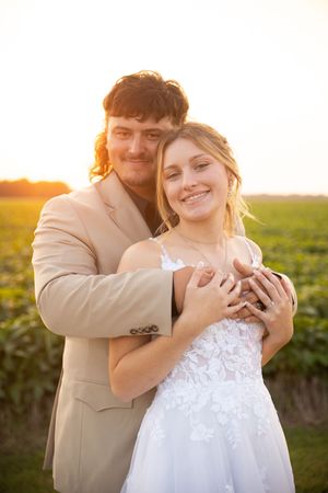 A couple embraces in a romantic sunset photoshoot against a green field backdrop wearing formal wedding attire.