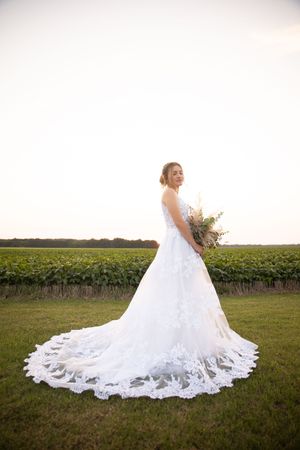 A stunning white wedding gown with a long lace train flows across a green field at sunset.