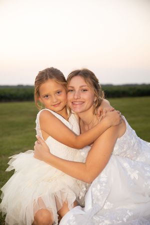 Two people in white dresses embrace outdoors during sunset in a grassy field.
