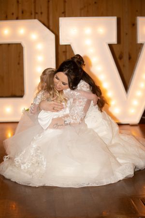 Two women in white wedding dresses share an emotional embrace in front of illuminated marquee letters.