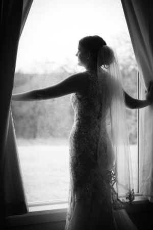 Silhouette of a bride in a lace wedding dress standing by a window with flowing curtains.