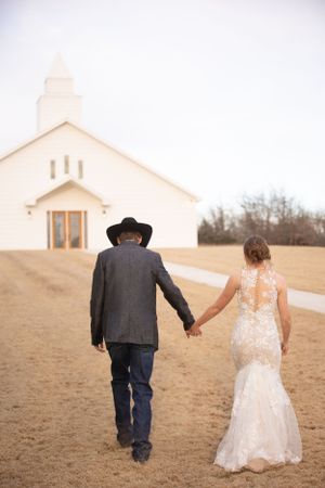Newlyweds walking hand in hand toward a white chapel in a rustic setting.