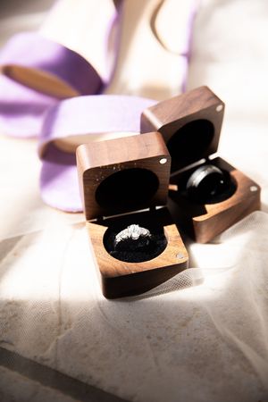 Two wooden ring boxes with a diamond engagement ring displayed inside on white fabric with purple ribbon in soft lighting.