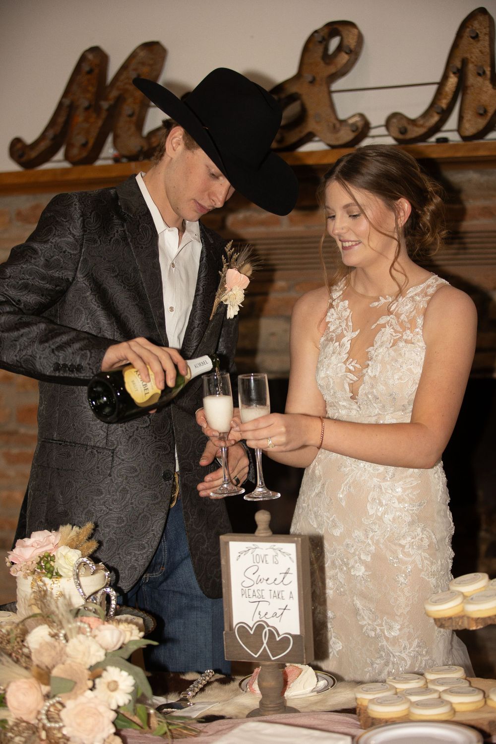 A couple pours champagne together at their rustic wedding reception with decorative decor in the background.