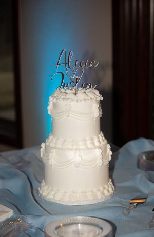 An elegant two-tier white wedding cake with a personalized topper and piped decorations.
