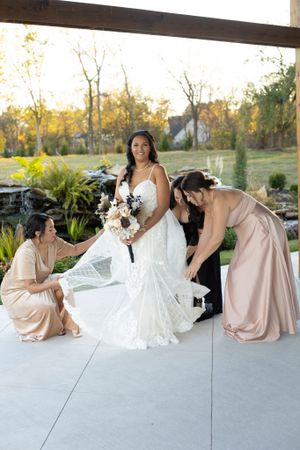 Bridesmaids in blush dresses helping adjust bride's wedding gown train outdoors.
