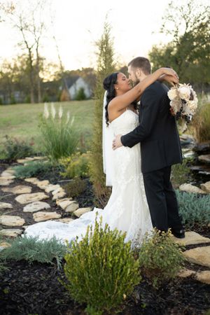 Newlyweds embrace on a garden path at sunset surrounded by ornamental grasses and landscaping.