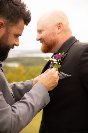 A groomsman helps adjust a purple and white floral boutonniere on another's suit jacket at an outdoor wedding.