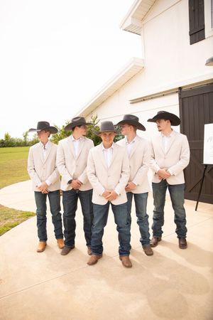 Groomsmen wearing cream blazers, cowboy hats and blue jeans pose together outside a white barn.