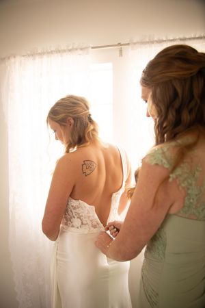 Person helps bride get dressed while standing near white curtained window in soft natural light.