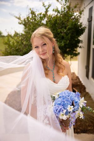 Bride in white dress and veil holds blue hydrangea bouquet while her veil flows in the breeze.