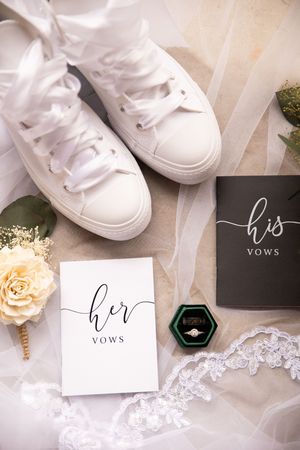 Elegant wedding details with white sneakers, vows cards, delicate lace veil, and peach roses on neutral background.