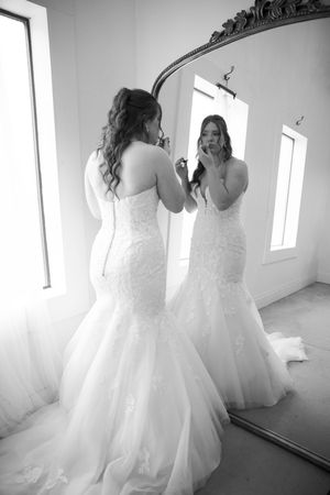 A person in a white mermaid-style wedding gown looks at their reflection in an ornate mirror.