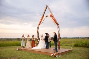 Rustic wooden wedding arch decorated with white fabric stands in an open field during a sunset ceremony.