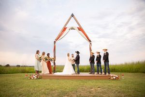 A wedding ceremony takes place under a wooden arch on a hilltop with wedding party members standing on both sides.