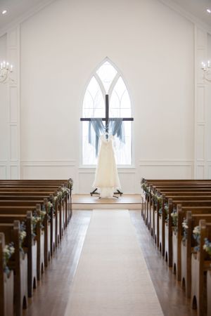 Elegant white wedding dress hanging in a church chapel with wooden pews and a gothic arched window.