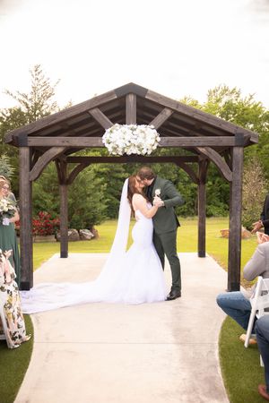 Romantic wedding ceremony under a rustic wooden pavilion decorated with white floral arrangements in an outdoor garden setting.