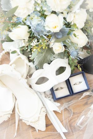 Wedding accessories including white floral bouquet, masquerade mask, and rings in navy boxes.