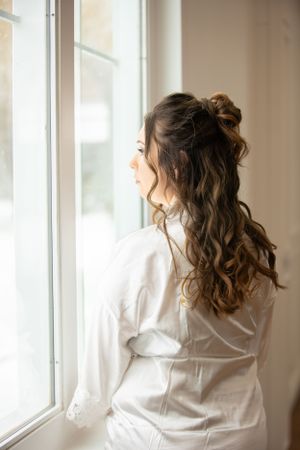Person in white robe gazing out window with styled curly hair for wedding preparation.