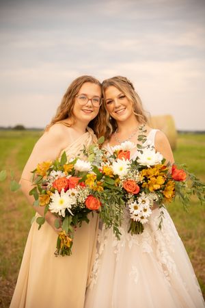 Two women in yellow bridesmaid dresses and white wedding gown hold vibrant floral bouquets in an outdoor setting.