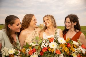 A group of bridesmaids in earth-toned dresses share a candid laughing moment while holding colorful autumn bouquets.