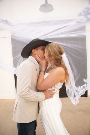 A bride and groom share their first kiss as newlyweds under a white veil with a rustic barn backdrop.