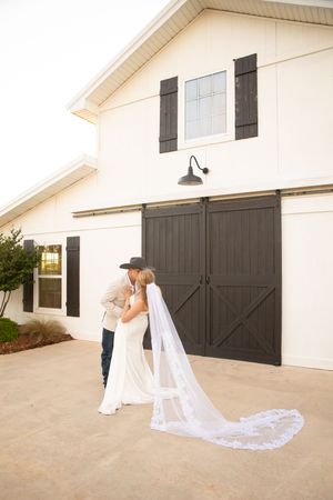 A white wedding dress train with intricate lace details flows gracefully on concrete against a modern farmhouse backdrop.