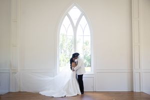 A bright white room with gothic arched windows and a couple in formal wedding attire sharing a tender moment.