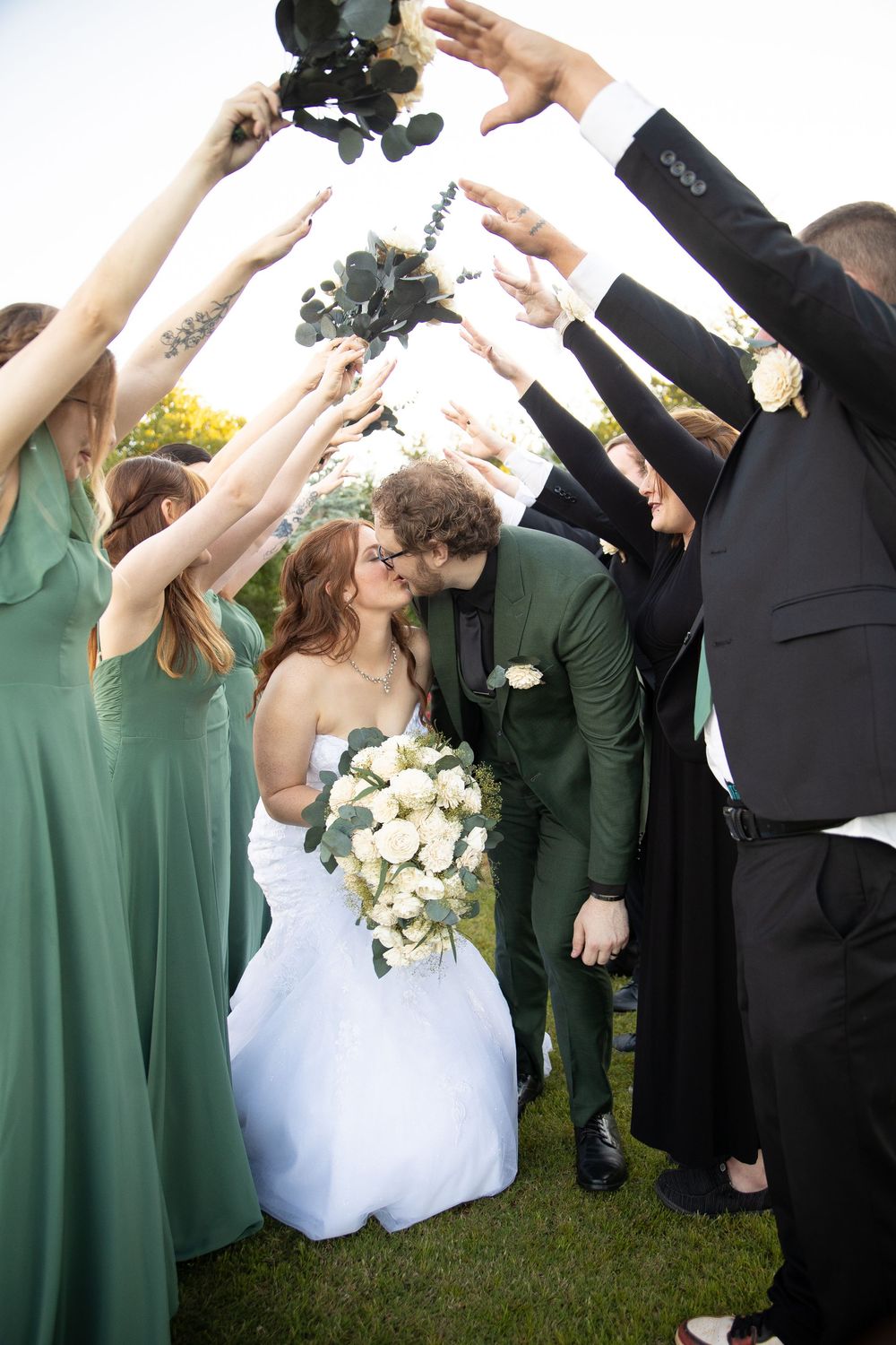 Wedding party members in green dresses and suits raise their arms during a celebratory moment with bouquets and flowers.