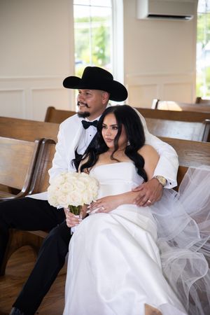 A romantic wedding couple in black and white attire relaxes together on wooden church pews.
