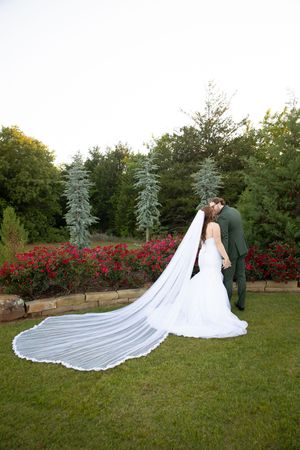 A couple in wedding attire share an intimate moment in a garden setting with flowing white veil and red flowers.