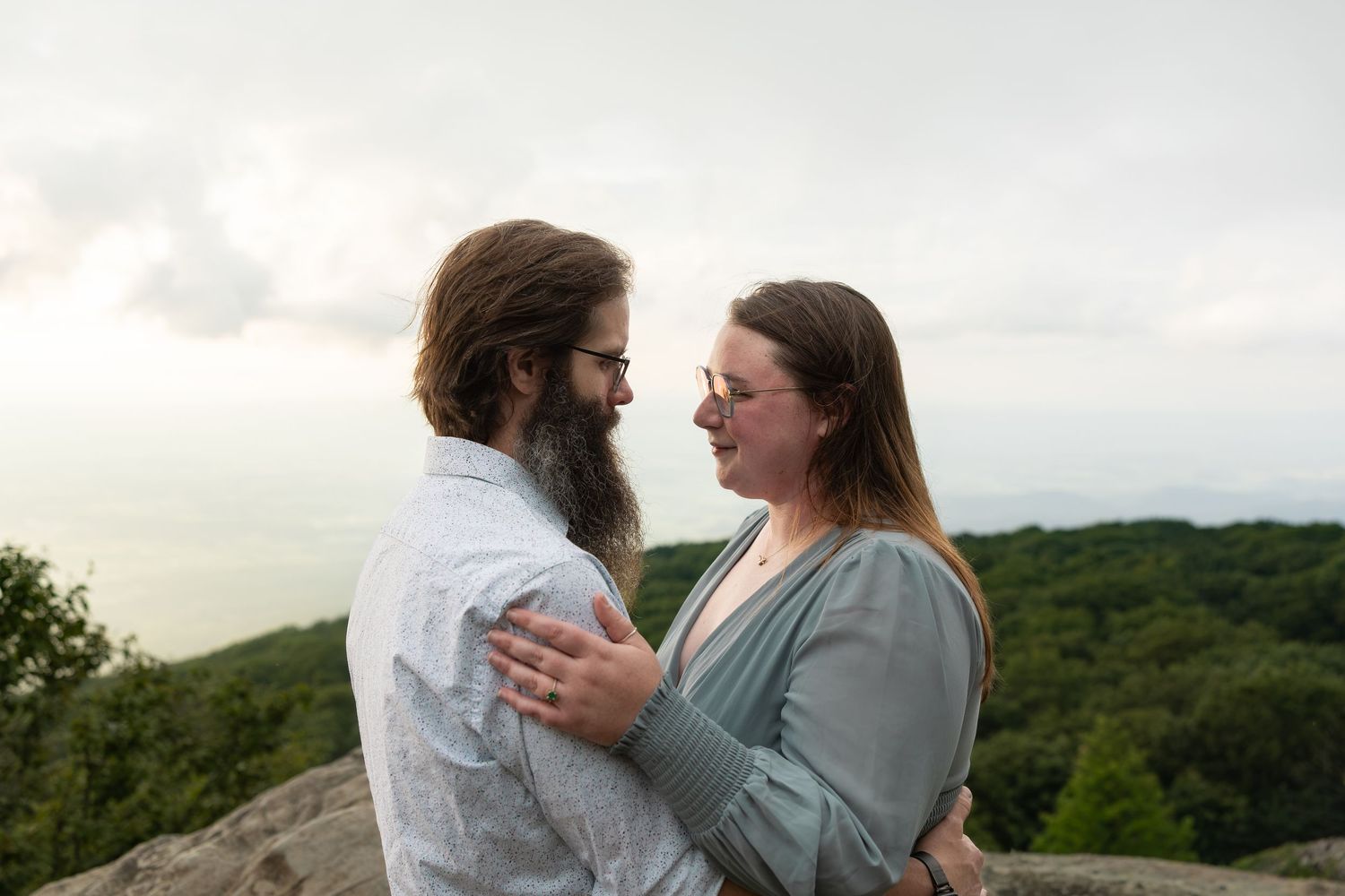 Two people standing on rocky cliff edge overlooking misty mountain landscape at sunset.