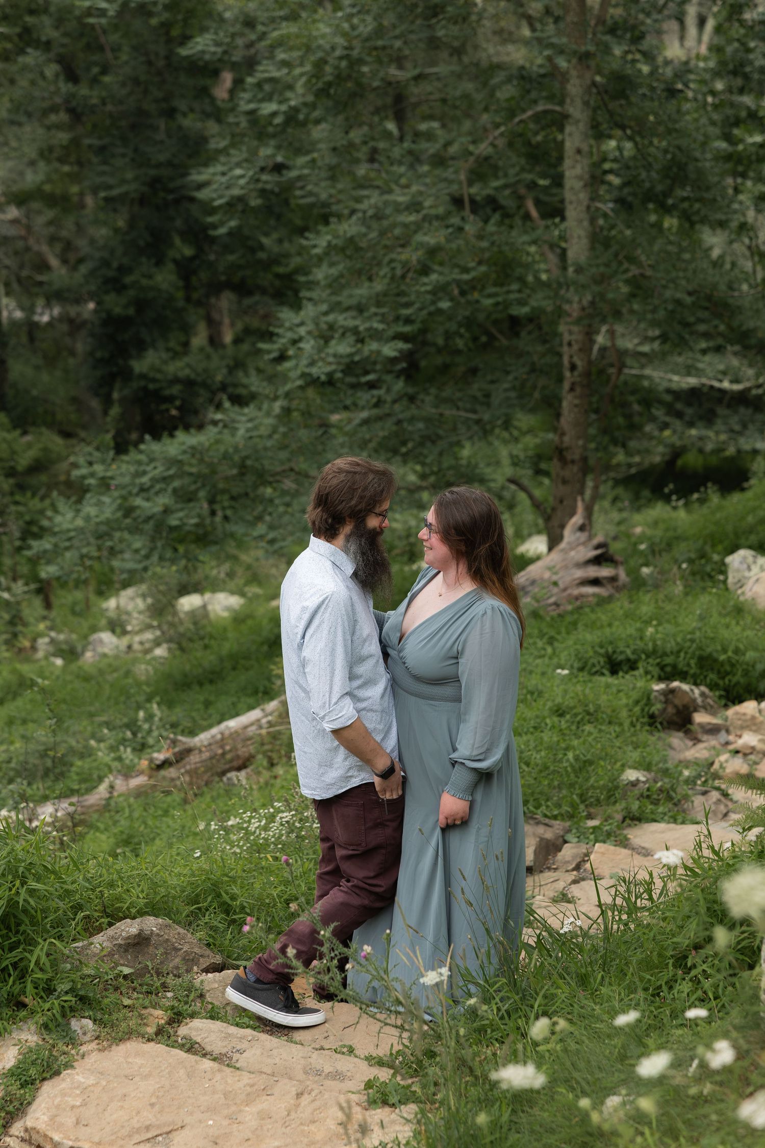 A joyful couple embraces while standing in a forest setting with bare trees and rocky terrain in the background.