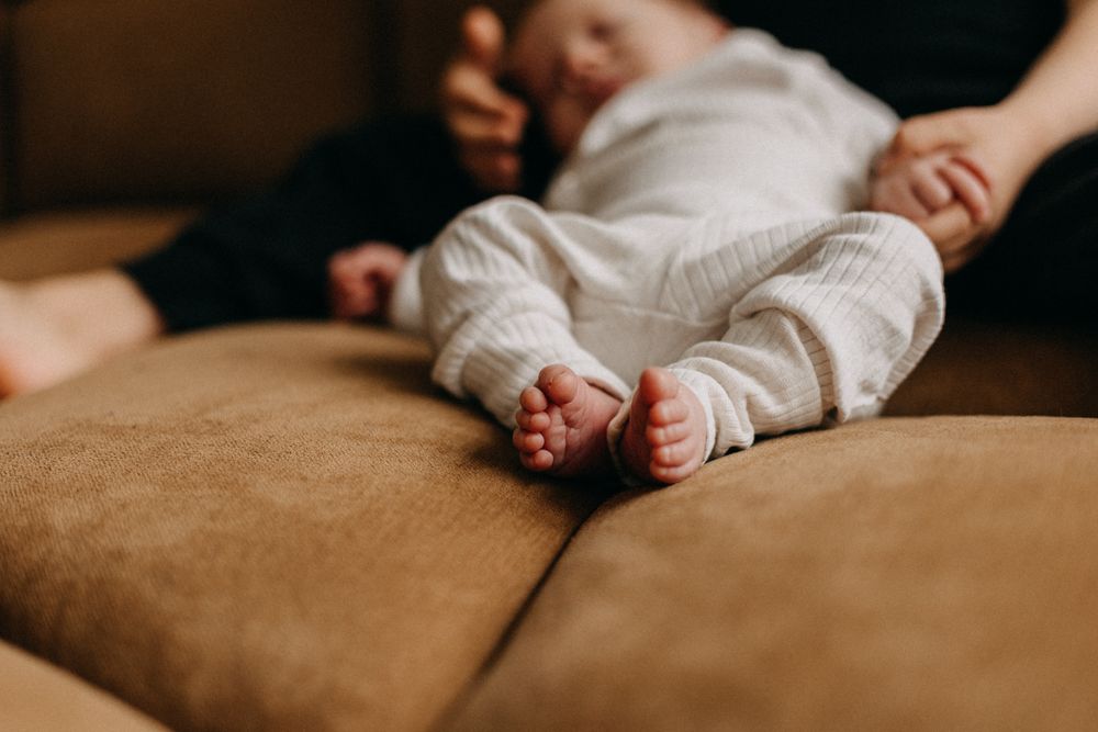 Close-up of baby feet in white onesie resting on brown couch cushion.