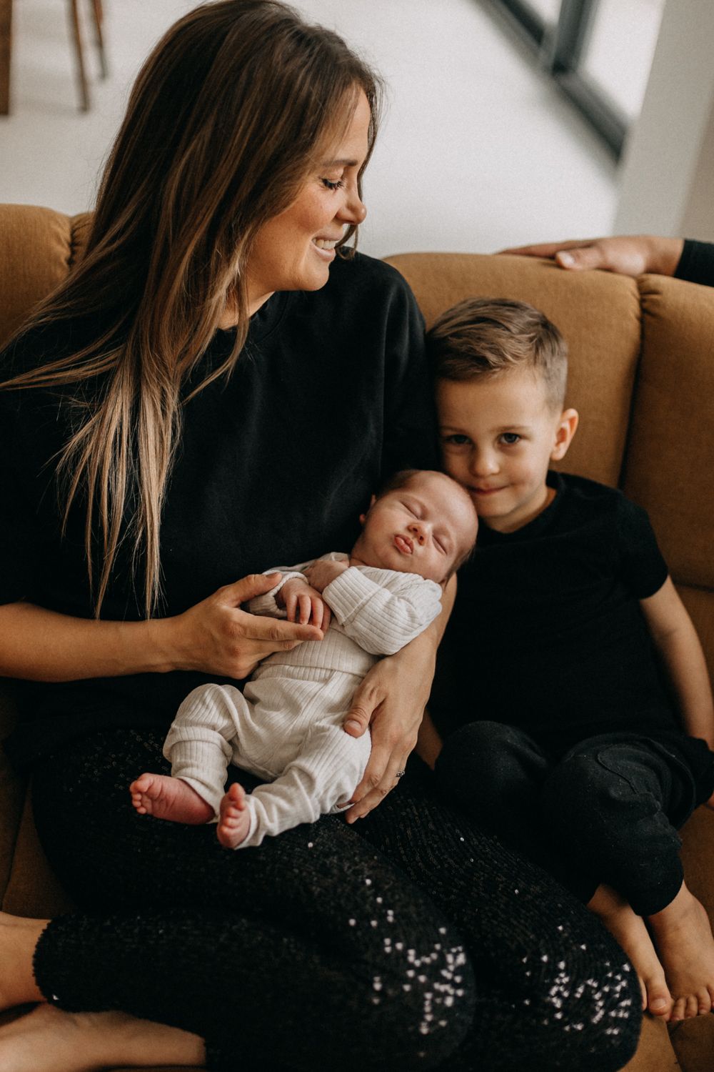 Family relaxing on brown couch with newborn baby and older child.