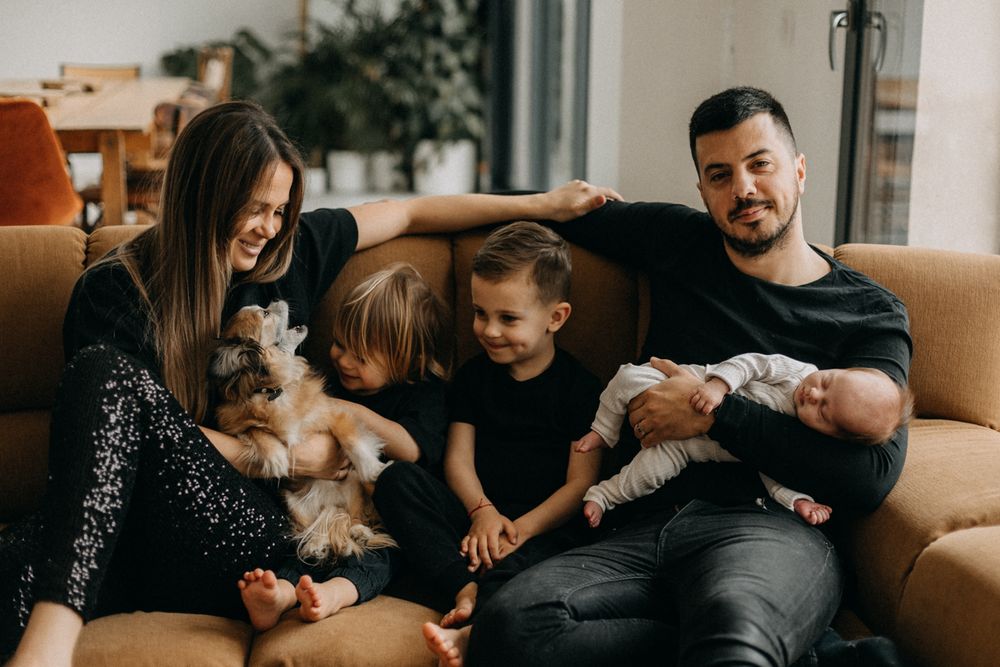 Family lounging on brown couch with small dog and infant in casual home setting.
