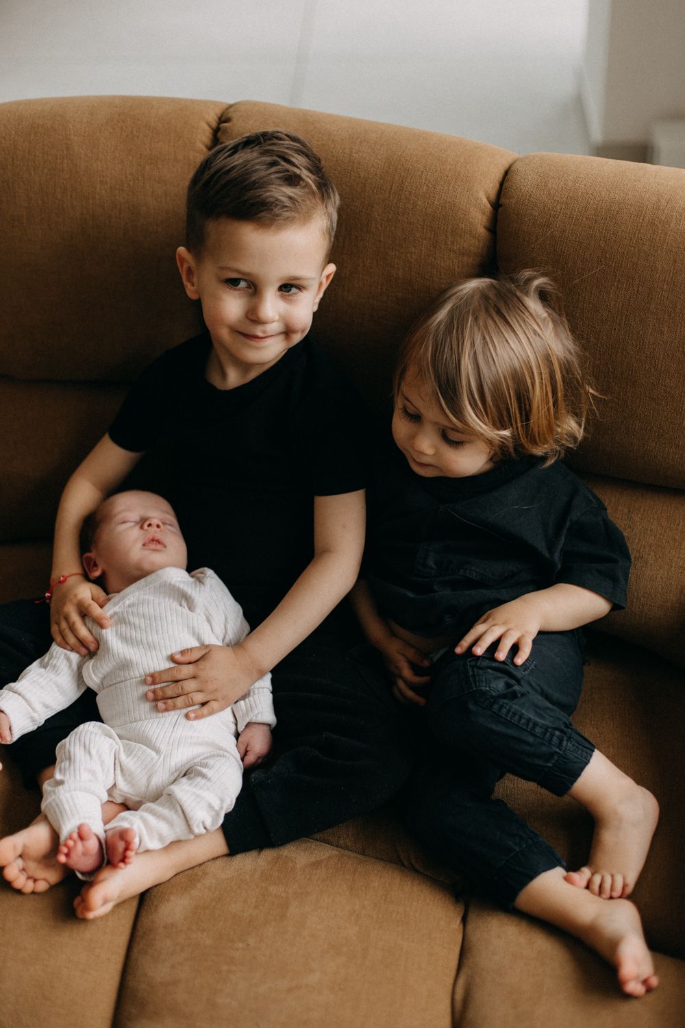 Three children sit together on a brown couch with the smallest in the middle.