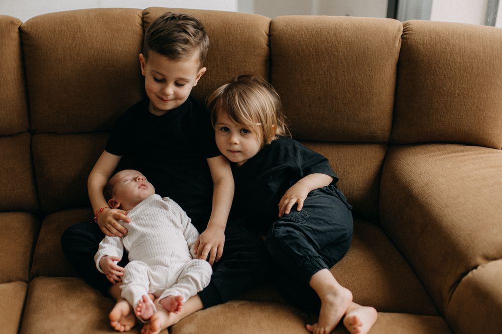Three siblings share a tender moment while sitting on a brown sofa.