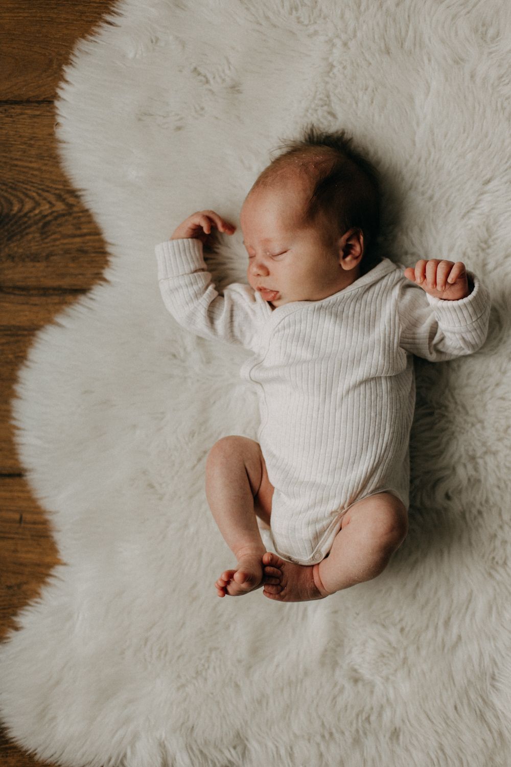 A sleeping infant in white ribbed clothing rests peacefully on a white furry rug.
