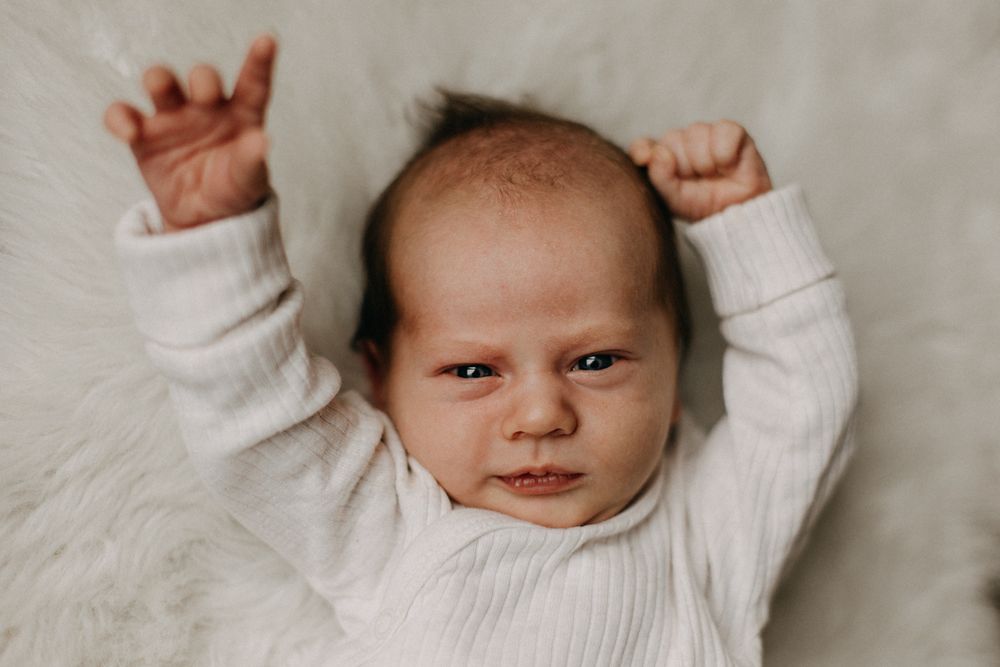 A smiling infant in white raises both arms while lying on plush white fabric.